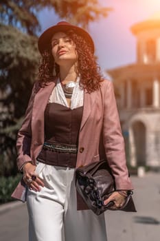 Woman park city. Stylish woman in a hat walks in a park in the city. Dressed in white corset trousers and a pink jacket with a bag in her hands