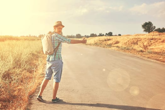 Hitchhiker with backpack in summer hat, light shirt, shorts on the road in tropical country in Sunny weather at autumn