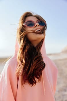 Stylish woman in sunglasses and pink sweatshirt enjoying windy day on beach with hair blowing