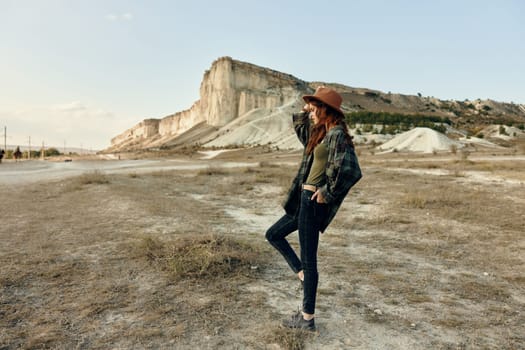 Woman in hat and black pants standing in front of hill with mountain backdrop on sunny day