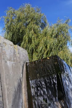 Fragment of a Waterfall against the background of a tree with green foliage on the river bank against a blue sky, view from below.