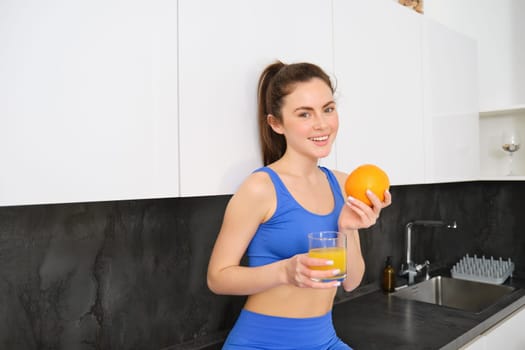 Wellbeing and sport. Young smiling nutritionist, fitness girl holding orange and fresh juice, drinking it from glass and looking happy, posing in sportsbra and leggings, standing in kitchen.