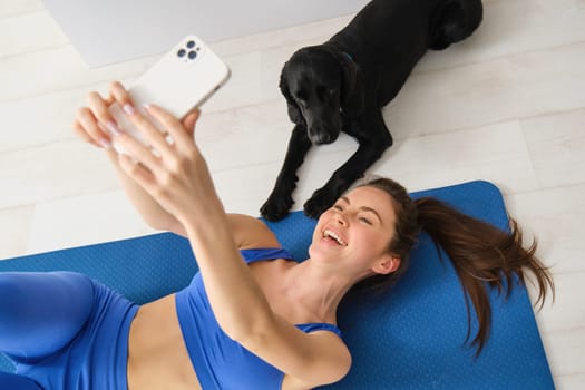 Portrait of happy young fitness woman, lying on mat and taking selfie with a dog in living room, workout with her pet.