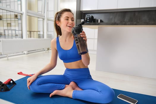 Young woman working out from home, drinking water during her sport training session in living room, sitting on yoga mat.