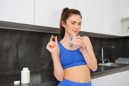 Portrait of healthy, smiling fitness woman, showing vitamins, fish oil dietary supplement for strong, fit body, drinking water with tablet, standing in ktichen.