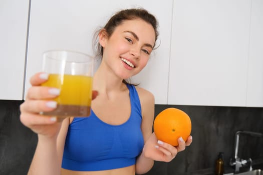 Portrait of smiling fitness woman, offering orange juice, holding fruit and a glass in hands, posing in kitchen in activewear.