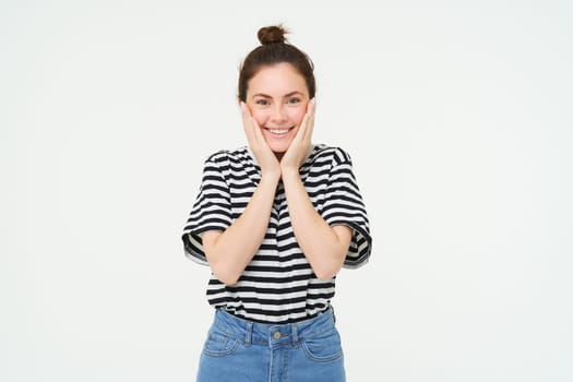 Portrait of cute smiling girl blushing, holds hands on cheeks and looks excited, receive happy news, stands over white background.