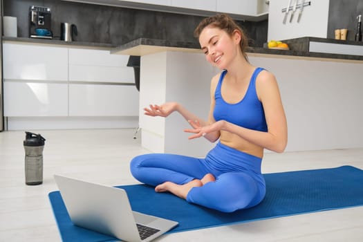 Image of young smiling woman, fitness instructor sitting on yoga mat, talking to client via online video chat on laptop, teaching aerobics, workout training.