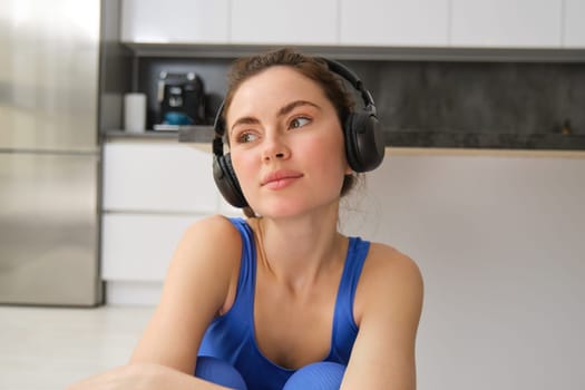 Beautiful brunette girl, doing fitness workout, sitting in living room in wireless headphones, listening to music and doing sports.