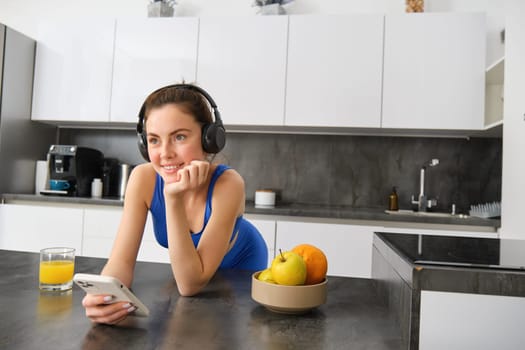 Lifestyle and workout. Young smiling woman in headphones, standing in kitchen with smartphone, drinking orange juice and listening music, heading over to gym, going jogging.