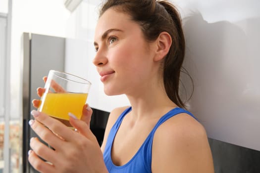Close up portrait of beautiful young woman, looking away, drinking orange juice, fresh in glass, standing in kitchen, wearing workout clothes.