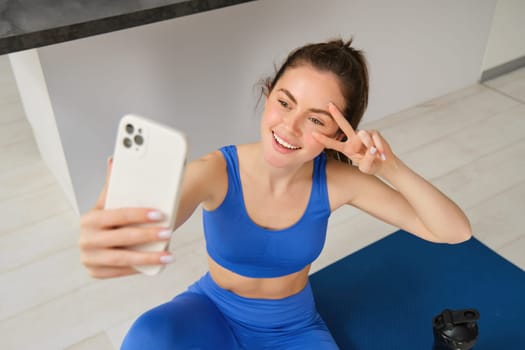 Portrait of beautiful female athlete, woman doing workout at home on rubber yoga mat, takes selfie on smartphone, makes photos for social media of fitness instructor.