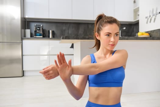 Image of fitness girl concentrates on workout, stretches hands before training session at home, follows online gym instructions.