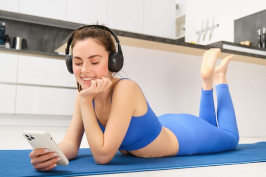 Portrait of fitness woman, listening yoga music in headphones, using her smartphone during workout at home, laying on blue sports mat.