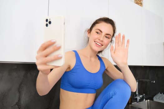 Portrait of sportswoman, fitness blogger, recording video, waving hand at smartphone screen and smiling, online chatting, sitting in kitchen, wearing activewear.