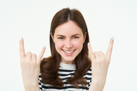 Close up portrait of excited young woman, rock n roll girl, showing heavy metal horn fingers and smiling, enjoys concert, having fun, standing isolated over white background.