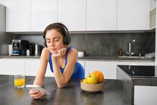 Image of happy, stylish young sports woman, standing in kitchen and drinking orange juice, listening music in headphones, using smartphone app.