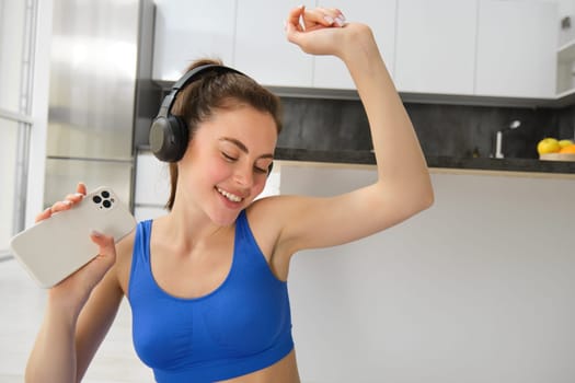 Portrait of happy, brunette woman in headphones, holding smartphone, dancing and working out, doing fitness exercises on yoga mat.