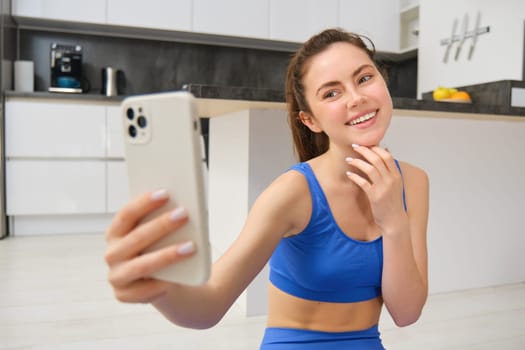 Portrait of young fitness girl, blogger records her workout from home, poses for selfie on smartphone, does yoga on camera, sits in blue sportsbra and leggings.