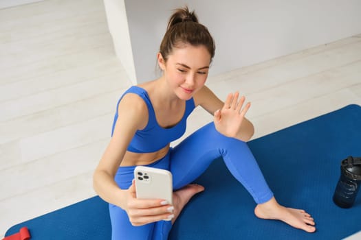 Image of beautiful, atheltic girl sits on yoga mat at home with smartphone, takes selfie and workout, does fitness exercises.