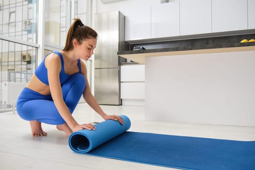 Image of young woman preparing living room for meditation, yoga exercises, unwrapping a mat on living room floor, wearing sportswear.