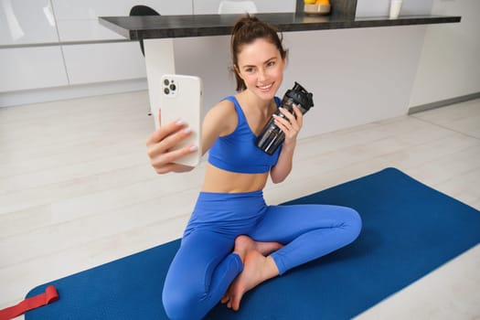 Portrait of woman doing sports, takes selfie on smartphone, fitness instructor records her exercises, stays hydrated, drinks water during training session.