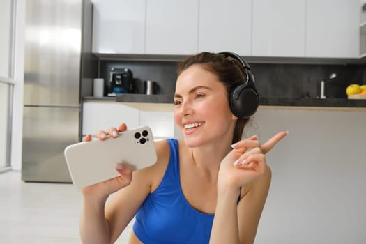 Close up portrait of young happy woman, workout at home, listens music in wireless headphones, singing at smartphone, doing training exercises.