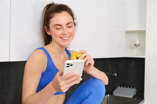 Healthy lifestyle and technology. Young beautiful girl, fitness instructor, looking at smartphone, reading on mobile phone and eating an apple, smiling happily.