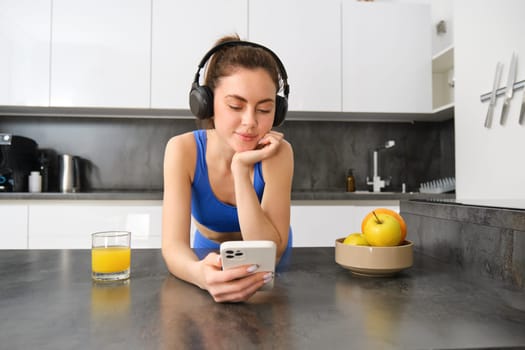 Portrait of young fitness woman with headphones, drinking orange juice in kitchen and using smartphone, listening music, getting ready for workout gym.