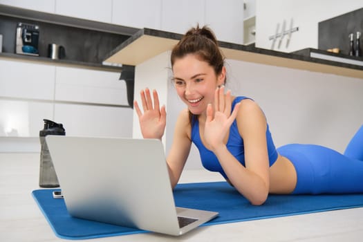 Portrait of beautiful, fit young woman on floor at home, saying hello at laptop screen, waving hand and smiling, workout remotely with friend, doing yoga class online.