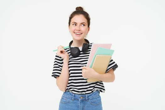 Portrait of young woman, student with notebooks and earphones on her neck, posing for college advertisement, white background.