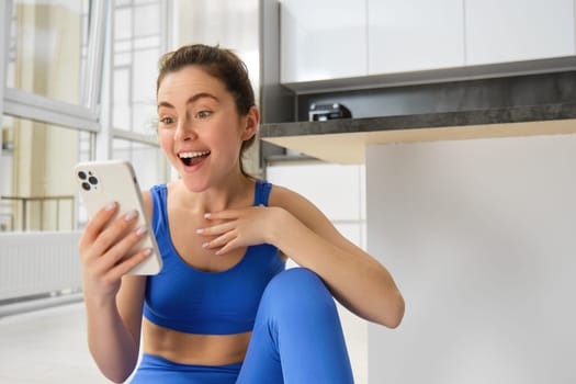 Indoor shot of surprised, happy young sportswoman looking at smartphone with smile and excitement, reading great news on phone message, sitting in blue sportsbra and leggings.