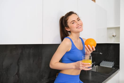 Indoor shot of woman after workout, standing in kitchen with fresh juice and an orange, drinking it.