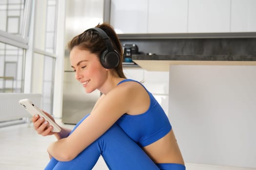 Portrait of brunette girl in fitness clothes, listens music in wireless headphones, holds smartphone, workout from home, looking at training video instructions.