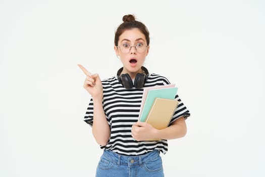 Portrait of girl gasps in surprised, points left with amazed face. Shocked student shows something, pointing finger at banner, holding notebooks, white background.