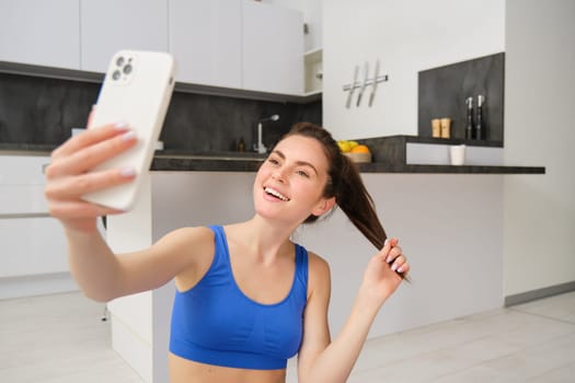 Close up portrait of young sporty woman, fitness girl takes selfie at home while doing her workout, posing on yoga mat, holds smartphone and smiles.