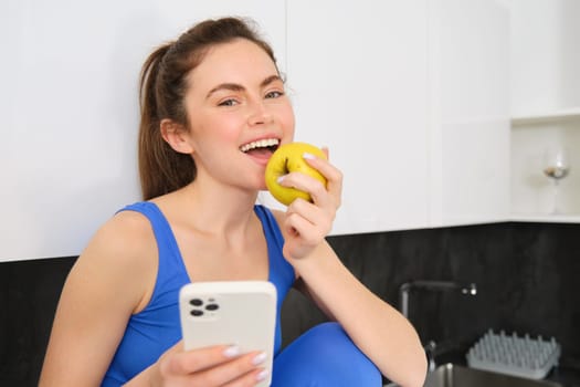 Portrait of smiling brunette girl, wearing fitness clothes, sitting in kitchen and biting an apple, eating fruit snack between workout, holding smartphone, using mobile phone app.