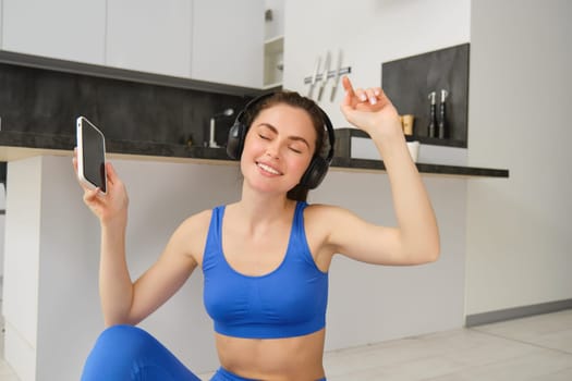 Cheerful brunette girl does fitness at home, wears wireless headphones and dances, holds smartphone, does workout indoors on floor, wears blue activewear.