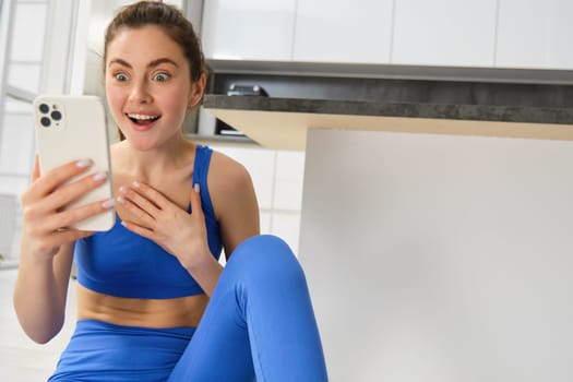 Close up shot of young happy woman, doing sports, workout at home, checking her phone, looking amazed at smartphone screen. Copy space
