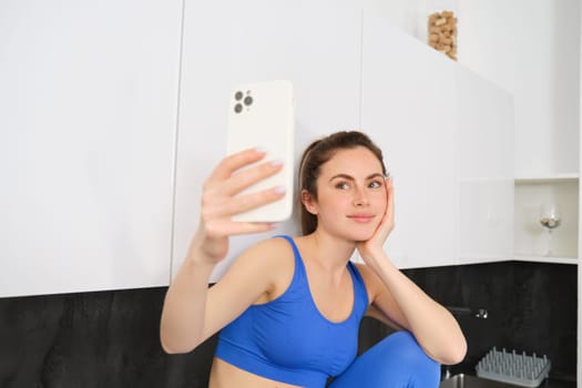 Portrait of fitness girl posing for photo, taking selfie on smartphone app, sitting in kitchen, wearing activewear.