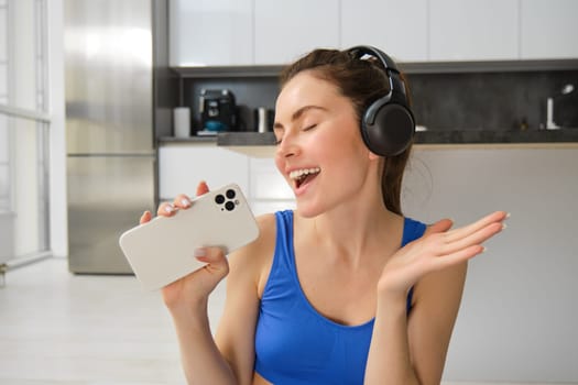 Close up portrait of young happy woman, workout at home, listens music in wireless headphones, singing at smartphone, doing training exercises.