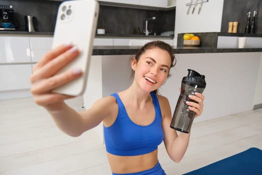 Portrait of sportswoman takes selfie with water bottle in living room, holds smartphone and poses for photo while doing fitness workout.