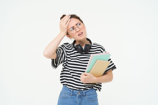 Portrait of compllicated, sad young woman in glasses, carries homework notebooks, facepalms, looks disappointed, stands over white background.