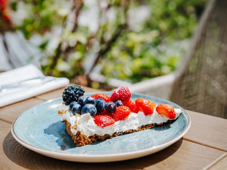 Piece of appetizing cheesecake or berry tart with whipped cream in blue rustic plate on wooden table outdoor in summer sunny day. New york cheesecake with berries mix, lifestyle shot