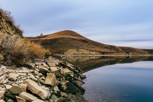 Big boulders on hilly coastline. Overcast sky reflected on lake surface.