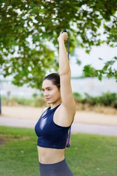 A young woman is in a park, using a resistance band for upper body exercises, focusing on strength training and wellbeing. Embracing fitness and fresh air, she engages in a workout