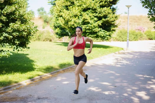 A young woman in sportswear jogs with determination in a sunny park, showcasing her dedication to fitness and a healthy lifestyle. Her commitment to wellness is evident in her energetic stride