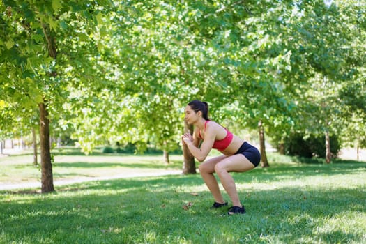 In a sunlit, green park, a young woman in athletic attire is deeply focused on her workout as she performs squats. The natural setting creates a serene atmosphere for her exercise