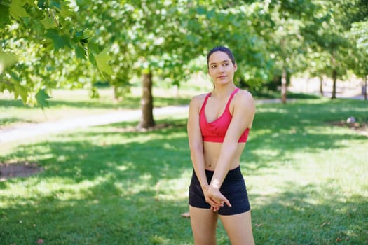 In a tranquil park, an athletic young woman is committed to her fitness and wellbeing, getting ready for an outdoor workout amid sunlit trees in nature. Her determination and focus are evident