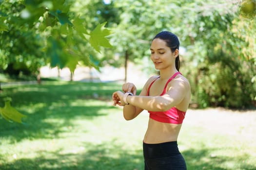 Young woman in red sports bra and black leggings checking her fitness tracker while exercising in a sunny, green park